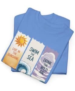 Light blue folded T-shirt displaying three vertical panels: an orange sun over wavy lines with “Live in the Sunshine,” a stylized wave with “Swim the Sea,” and a sky-and-wave scene with “Drink the Wild Air.