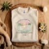 Sand colored flat fold Children Are a Gift From God Floral Christian T-Shirt featuring a watercolor sunrise, handprint flowers, and faith-based quote.